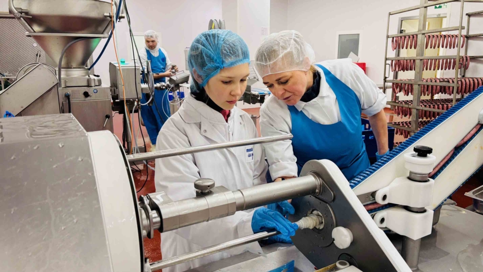 Mädchen mit Schutzhaube und Mitarbeiterin an Wurstmaschine